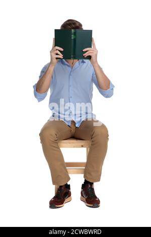 Young casual man covering his face with a book while sitting on a chair on white studio background Stock Photo