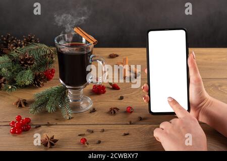 Female hand is holding cell mobile phone on the Christmas winter decorations. mulled wine with cinnamon. Modern cellphone with white blank screen in w Stock Photo