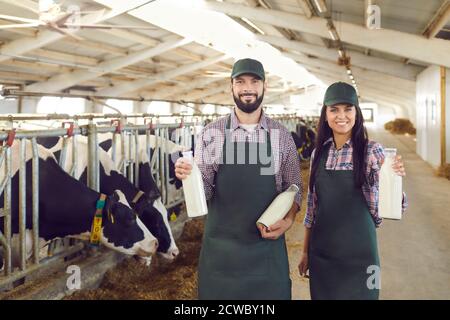 Happy dairy farm owners standing in cowshed, holding bottles of milk, smiling and looking at camera