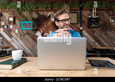 young casual man wearing glasses sitting at desk with hands together working on laptop and looking aside happy at the coffeeshop Stock Photo