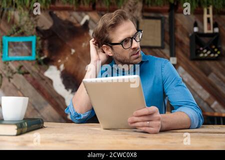 casual man wearing glasses sitting at desk and holding his tablet while scratching head and looking aside pensive at the coffeeshop Stock Photo
