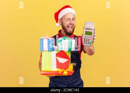 Happy positive courier in santa claus hat holding many gift boxes and showing payment terminal winking at camera, asking to pay contactless. Indoor st Stock Photo