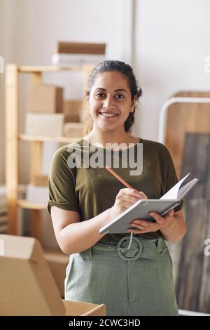 Portrait of young woman smiling at camera while making notes in note pad working in warehouse Stock Photo