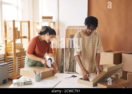 Two colleagues packing new clothes into the cardboard boxes at the table in warehouse they preparing parcels for delivery Stock Photo