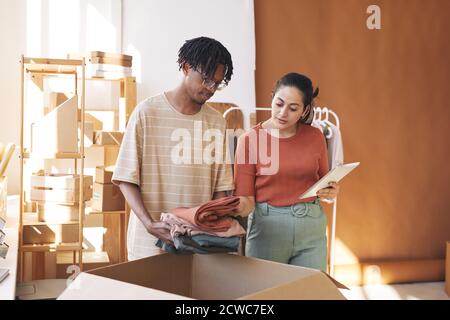 Woman using digital tablet she checking the orders while African man putting clothes into the big cardboard box for delivery Stock Photo