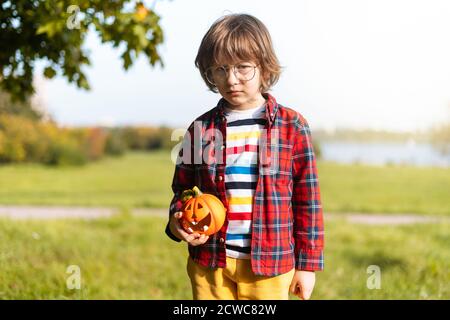 Cute sad boy in glasses play with pumpkin in autumn park on Halloween. Kids trick or treat. Boy carving pumpkins. Fun in fall. Dressed up child. Stock Photo