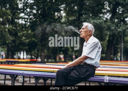 Senior adult men smoking cigarette outdoors in the city  park when sitting on the bench Stock Photo