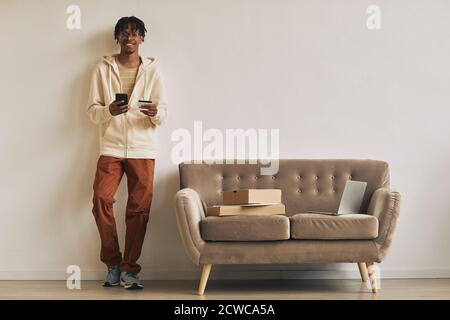 African young man leaning on the wall using his mobile phone with laptop and parcels on the sofa he getting his orders Stock Photo