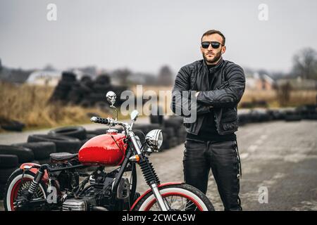 Red motorbike with rider. A man in a black leather jacket and pants stands near a motorcycle with his hands clasped, in the road. Tires are laid on th Stock Photo