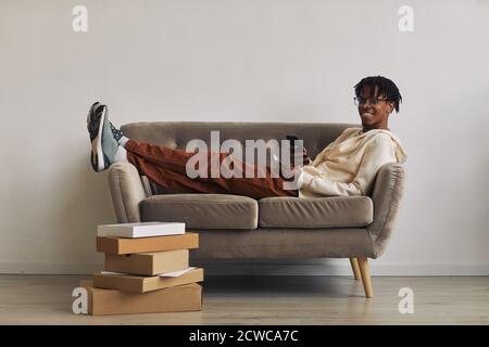 Happy African man lying on sofa with stacks of parcels on the floor and smiling at camera while buying purchases online on the phone Stock Photo