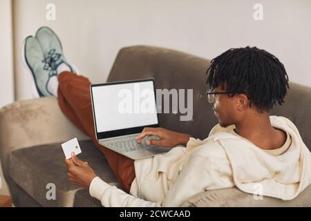 Rear view of African man lying on sofa with laptop he working online and using credit card for paying the bills Stock Photo