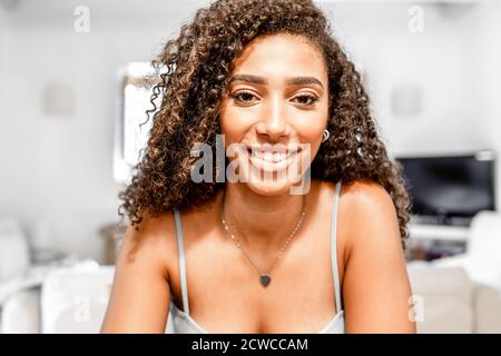 Close-up of young hispanic cute curly woman smiling looking at the camera like as in video conference in a white home living room ambient - Female con Stock Photo