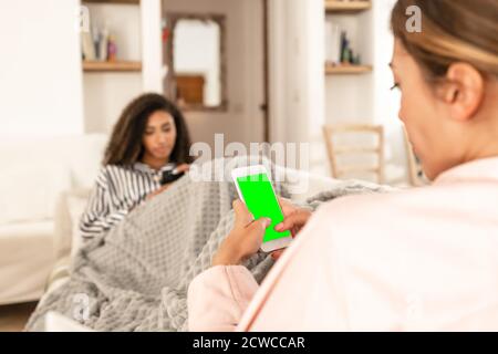 Two young mixed race female friends always connected communicating wearing pajama snuggle up on the sofa under a blanket - Selective focus on the gree Stock Photo