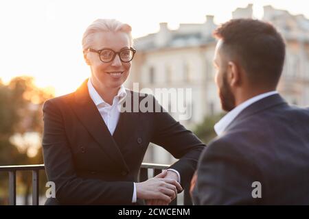 Mature businesswoman in eyeglasses and in suit smiling while talking to businessman at balcony during sunset in the background Stock Photo