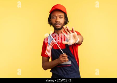 Serious afro-american craftsman with dreadlocks in uniform holding us flag showing stop gesture raising arm, no to racism, protection of human rights. Stock Photo