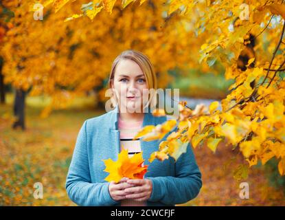 Beautiful blonde woman in autumn park holding maple leafs in her hands. Female outdoor fall portrait. Stock Photo