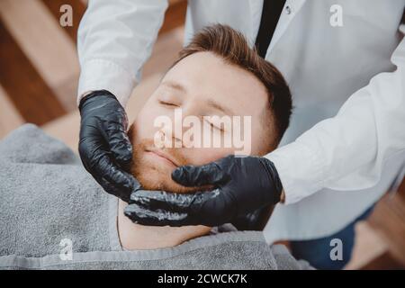 Barber hairdresser applies foam and beard shaving lotions to male client. Cream and oil skin care concept Stock Photo
