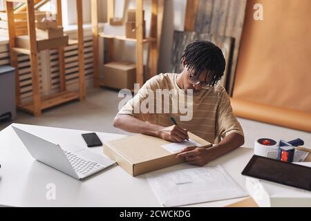 African young man sitting at the table in front of the laptop and writing address on the box before shipment Stock Photo