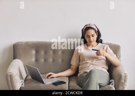 Young woman looking at number of her credit card while sitting on sofa and using laptop for online shopping Stock Photo