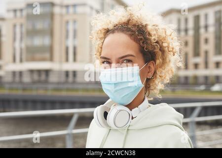 Portrait of young woman with curly hair wearing protective mask and looking at camera while walking in the city Stock Photo