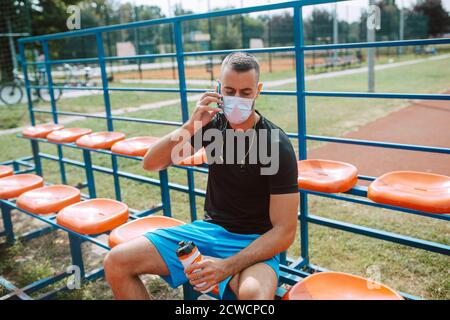 A fit young male caucasian athlete with a face mask sitting on the stands of a football field and talking on the phone. COVID - 19 coronavirus protect Stock Photo