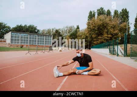 Sporty muscular caucasian male athlete with face mask looking away, stretching legs at the stadium race track. Muscular athlete outdoors.  COVID - 19 Stock Photo