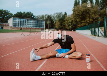 Sporty muscular caucasian male athlete with face mask looking away, stretching legs at the stadium race track. Muscular athlete outdoors.  COVID - 19 Stock Photo