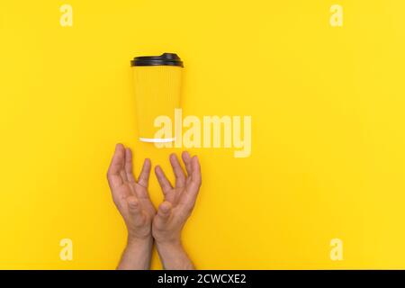 Two caucasian male hands and yellow paper cup with black lid with coffee take away on yellow background. Stock Photo