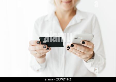 Unrecognizable woman wearing white shirt holding black credit card and smartphone, making online payment, online shopping, lifestyle technology Stock Photo