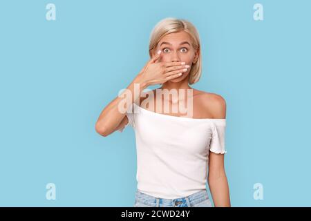 Freestyle. Young blonde woman in top isolated on blue covering mouth shocked Stock Photo