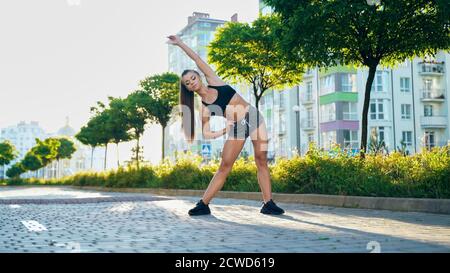 Front view of muscular young woman bending aside with hand on waist in sunny morning. Charming girl practicing relaxing exercises, warming up upper body in city streets near multistorey buildings. Stock Photo