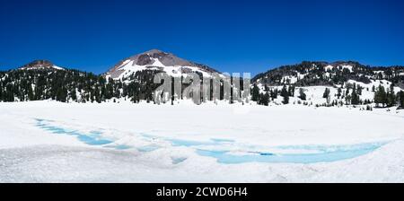 Forest and mountain at the back of lake Helen, covered in ice and snow in July on a sunny day with a mountain blue sky. Lassen National Park. Stock Photo