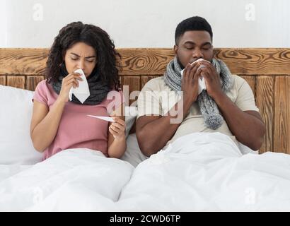 Sick african couple sitting in bed, measuring fever, sneezing noses Stock Photo