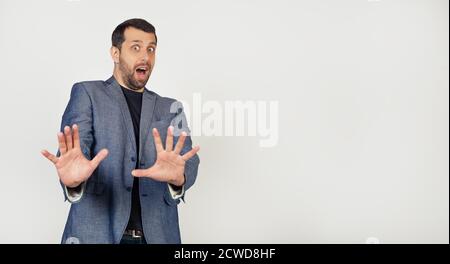 A young businessman with a smile, a man with a beard in a jacket, frightenedly stretched his arms forward with a stop gesture, with an expression of fear, screaming in shock. Portrait of a man. Stock Photo