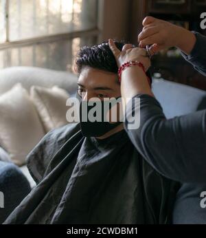 A man getting his hair cut at home by his mother. Young black haired latin wearing a mask, sitting at the living room. Stock Photo