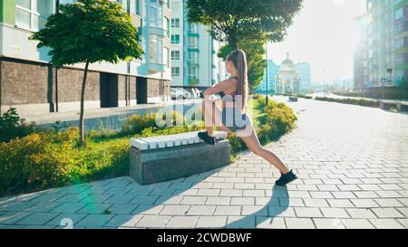 Unrecognizable fitness woman stretching leg using bench, doing flexibility exercises after running workout in city streets. Young woman warming up in rays of morning sun before training. Stock Photo