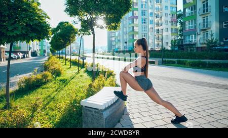 Side view of fitness woman wearing sportswear stretching leg on bench, practicing flexibility exercises after running workout in city streets. Stunning fit woman warming up in rays of morning sun. Stock Photo