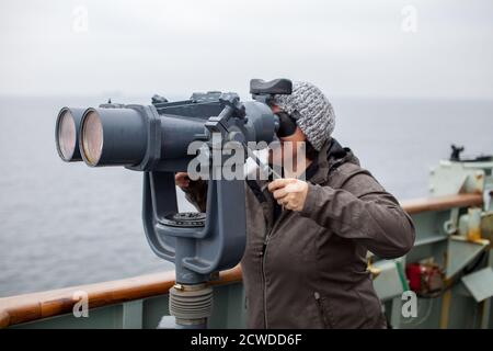 A young woman looks out through ship binoculars out into the open ocean at the Canadian Forces Base in Esquimalt, British Columbia, Canada Stock Photo