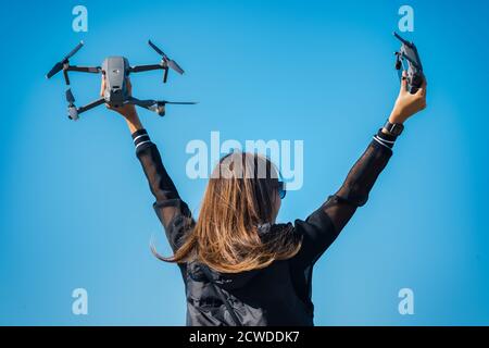 Young woman in black jacket holding remote control and drone before the flight on the blue sky background. Flying drone outside in summer. Stock Photo