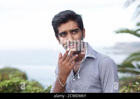 Happy ethnic man smoking cigarette, smiling, thinking looking sidewards outdoors background. Stock Photo