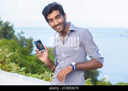 Portrait closeup of man showing phone screen smiling happy. Mixed race Indian Caucasian Latin model standing outside green bush and blue sea on backgr Stock Photo