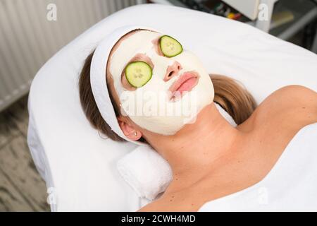 Woman lay on couch under towel with white alginate professional peeling clay mask and round cucumber slices cover eyes, cosmetologic skincare concept Stock Photo