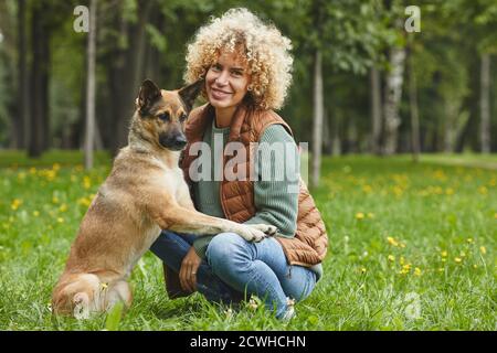 Portrait of young woman with blond curly hair smiling at camera while sitting on green grass with dog Stock Photo