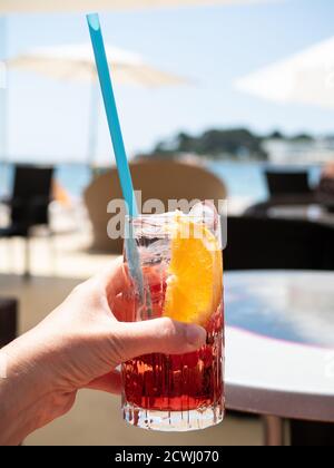Female hand holding glass of campari with soda and plastic straw. Beach with sunshades in sunny day. Stock Photo