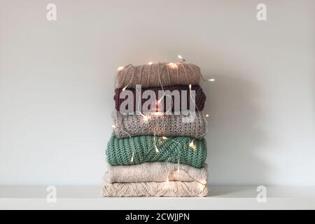 Stack of warm knitted women's sweaters on white background. Garland lights as decoration. Winter holidays concept. Stock Photo
