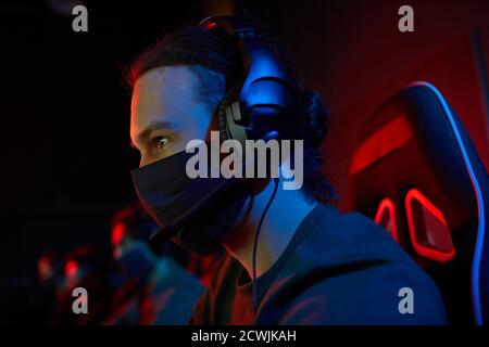 Young gamer in headphones and in protective mask playing computer games in dark room Stock Photo