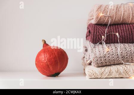 Pumpkin and stack of warm knitted women's sweaters on white background with garland lights. Winter holidays concept. Stock Photo
