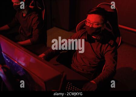 Hacker in protective mask sitting on chair in front of computer monitor working with new software in dark office Stock Photo