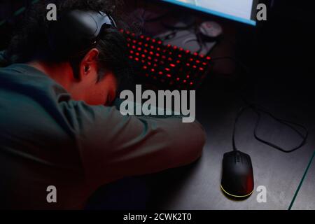 Rear view of young gamer in headphones sleeping at the table during game in dark room Stock Photo