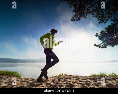 Slim man jogging on the beach early morning. Runner in yellow black running suit during run along lake. Stock Photo